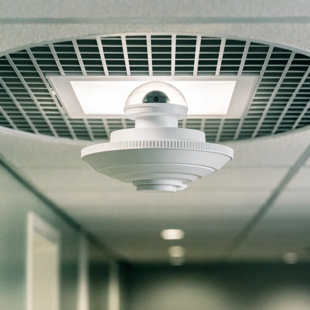 Vue rapprochée d'un détecteur de présence intégré au plafond d'un couloir de bureau, panneau LED rectangulaire allumé visible en arrière-plan, ambiance de lumière neutre 4000K